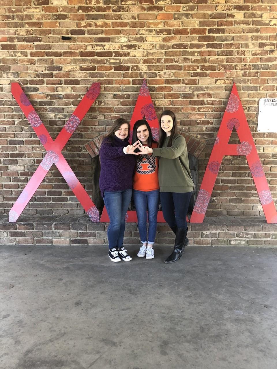 Members of Alpha Delta Chi in front of large letters throwing the organization sign with their hands. 